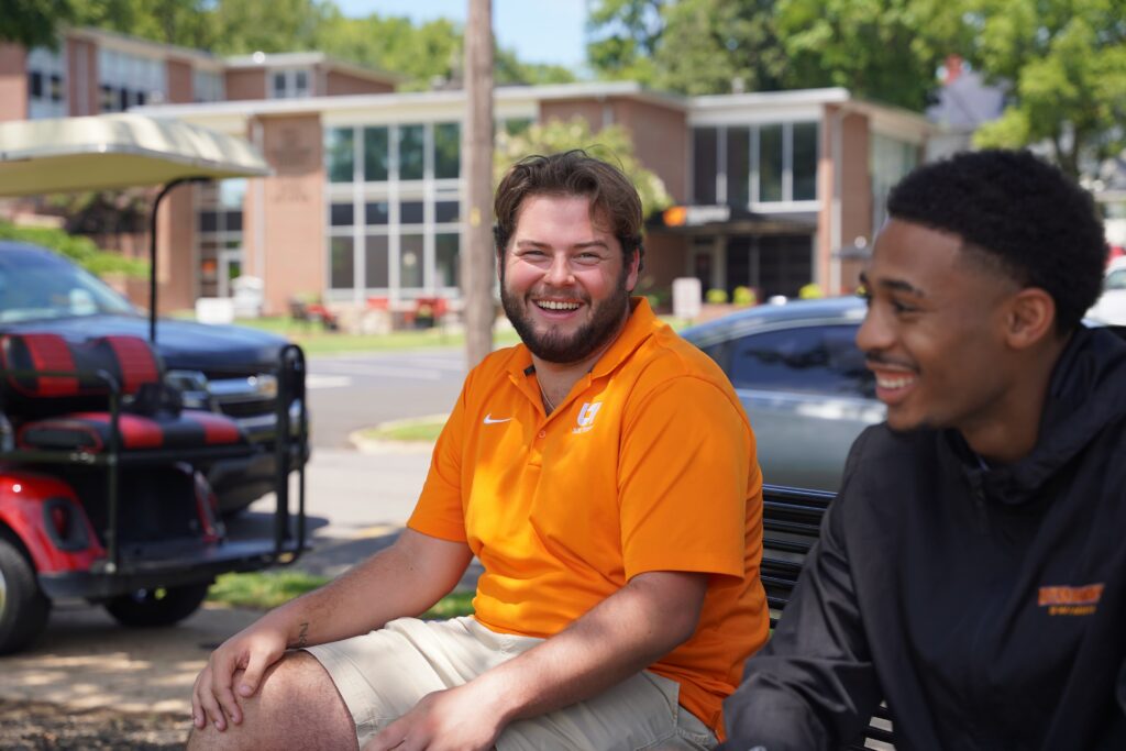 A student in orange smiling at the camera, with another smiling student in the foreground