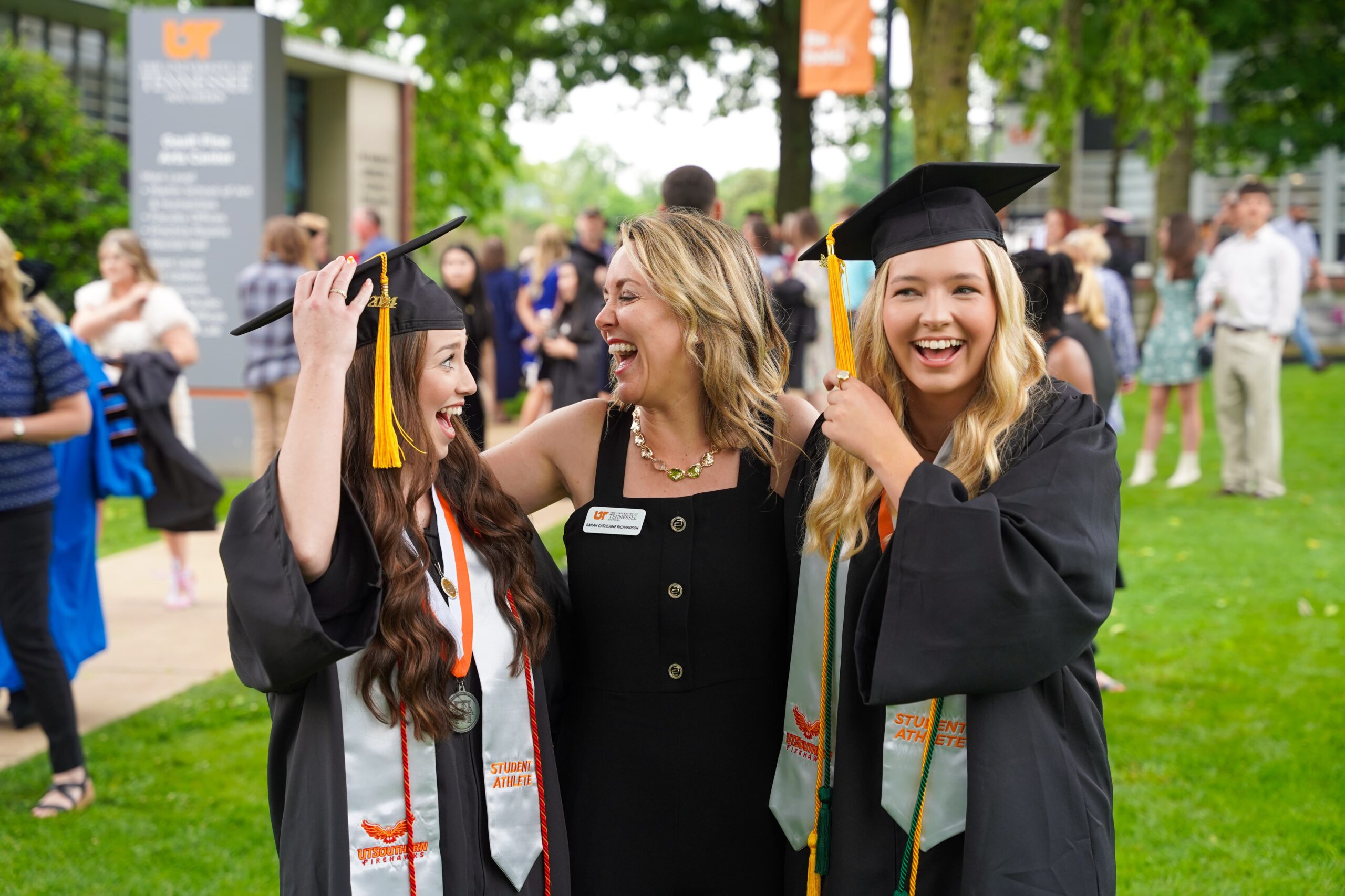 Two graduating students in caps and gowns with a faculty member in the middle, smiling