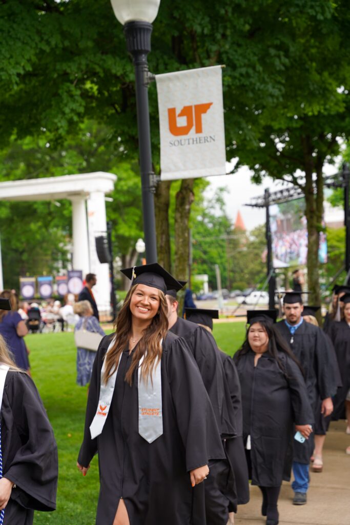 A student in cap and gown processing, smiling at the camera