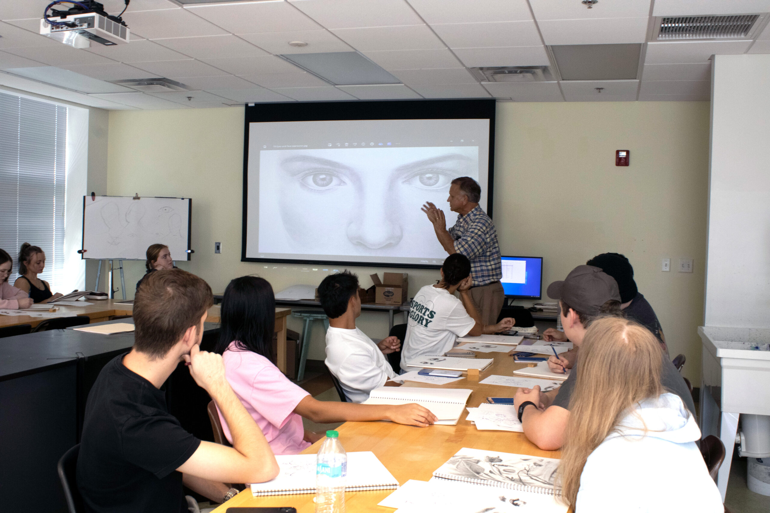 Classroom of students looking towards front of room. The instructor is showing a drawing of a face.