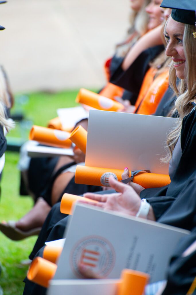 Students sitting in a row at graduation in cap and gown holding a program.