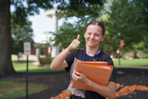 Female student holding her hand up with thumbs up and smiling.