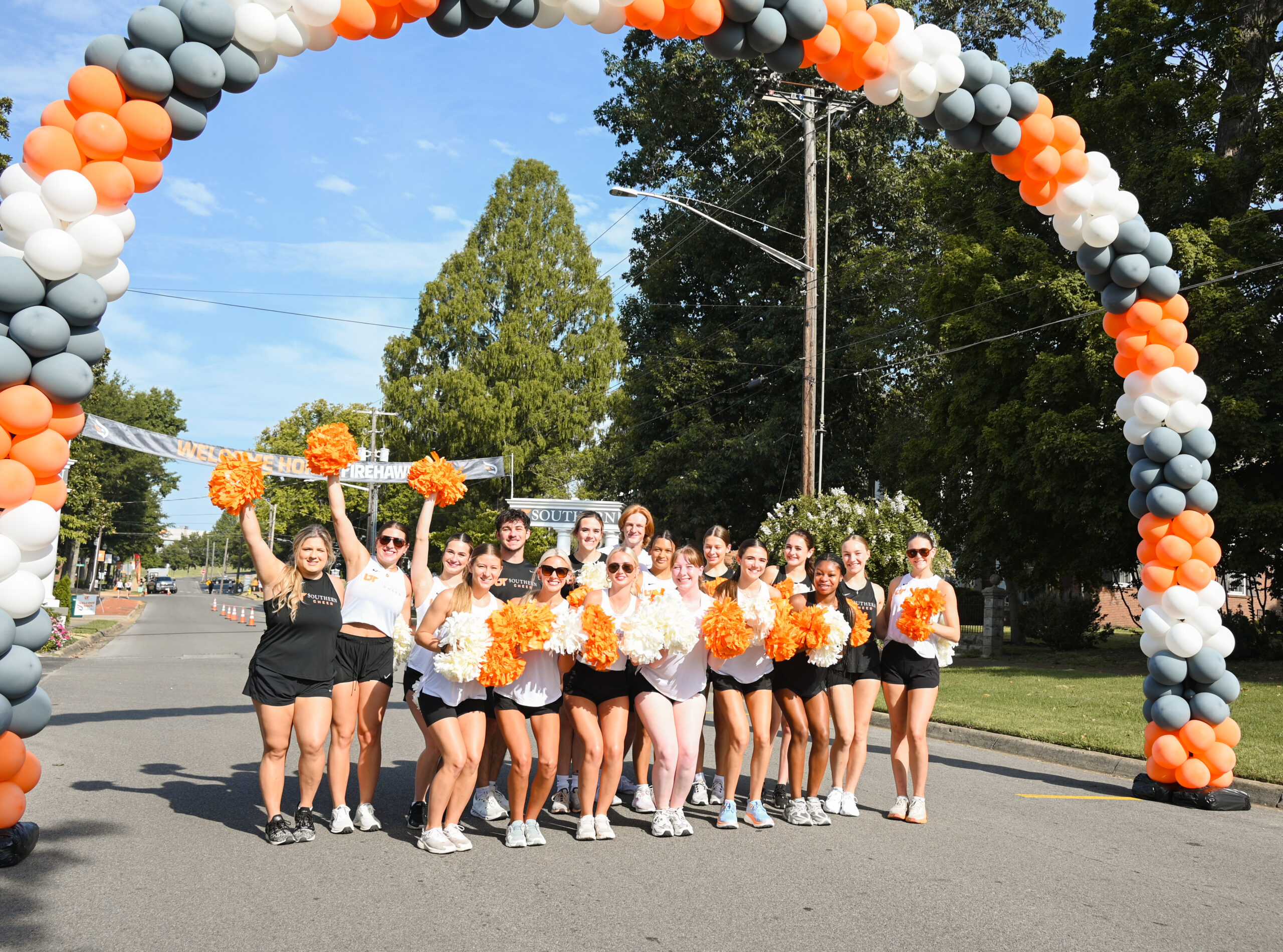 Cheerleaders gather and cheer under a orange, white and gray balloon arch that stretches over the street.