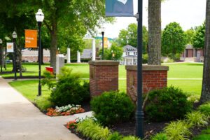 Scenic view of the University of Tennessee Southern campus featuring manicured lawns, flower beds, brick columns with plaques, and UT Southern light pole banners reading “Think big,” framed by trees and historic campus buildings in the background.